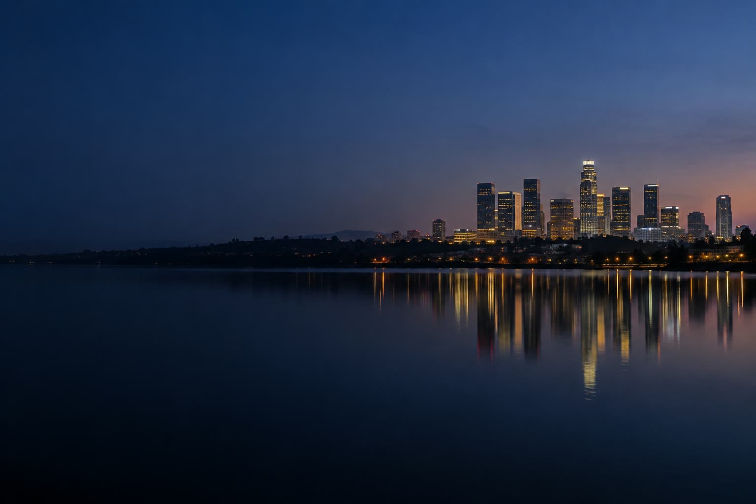 Los Angeles at blue hour, downtown towers small on the right horizon, with still dark water filling the left of the frame.