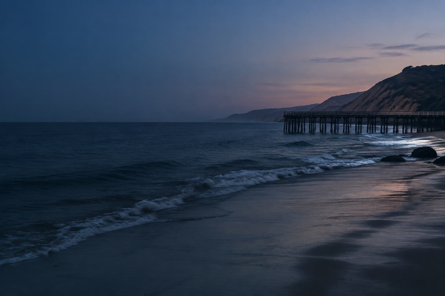 The Malibu coast at blue hour, a wooden pier and bluffs on the right, with dark Pacific water filling the left.