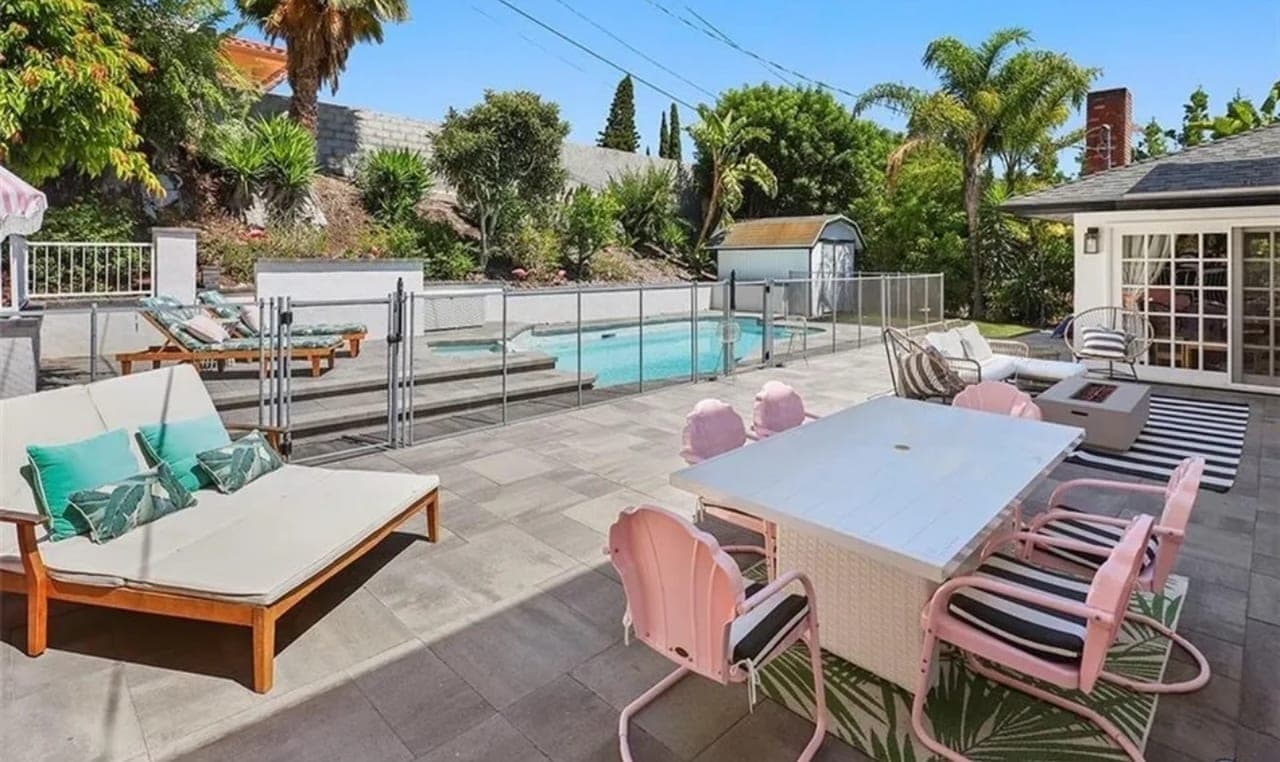A paved pool deck with loungers, an outdoor dining table and planting along the fence.