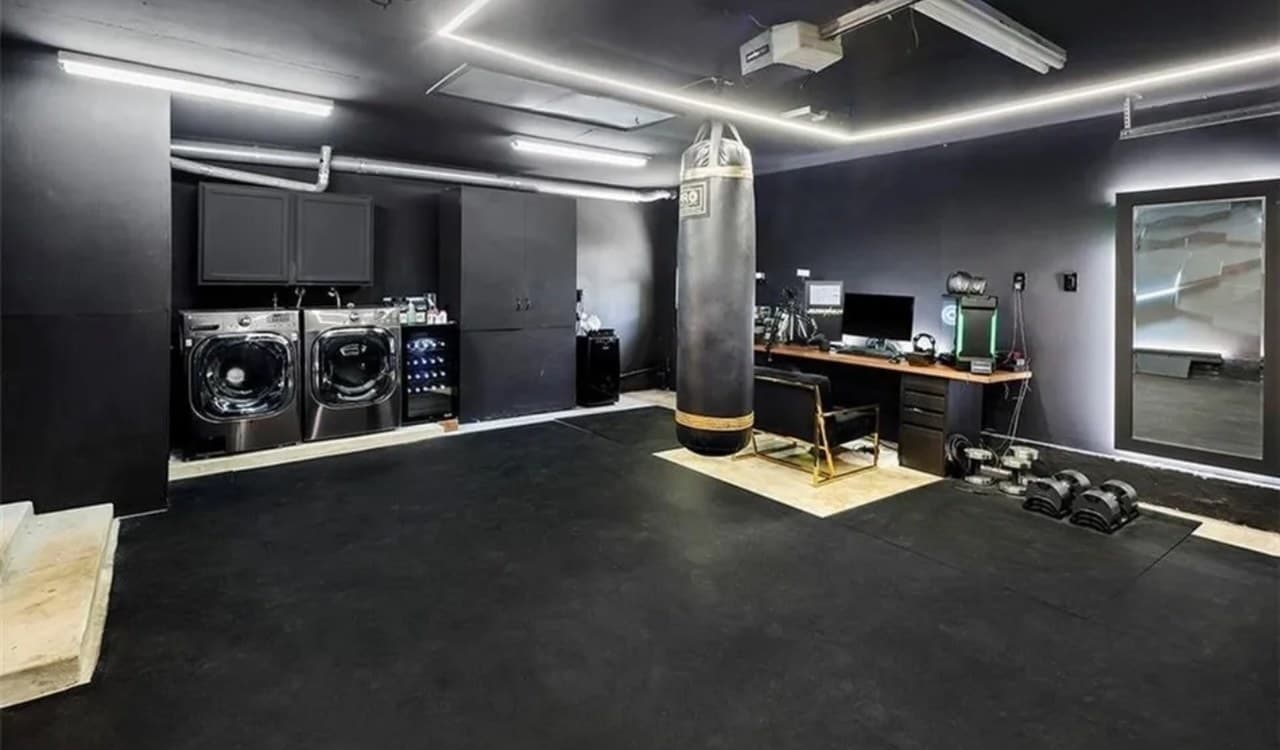 A converted garage gym with black walls, a hanging punching bag, dumbbells on a rubber floor, a mirror and a desk.