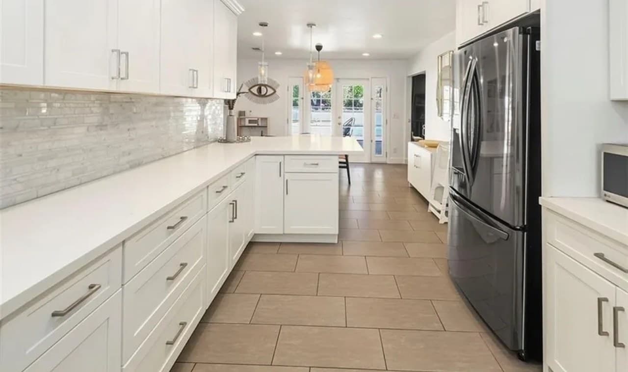 A long white kitchen with shaker cabinets, a dark refrigerator and a tiled floor running to French doors.