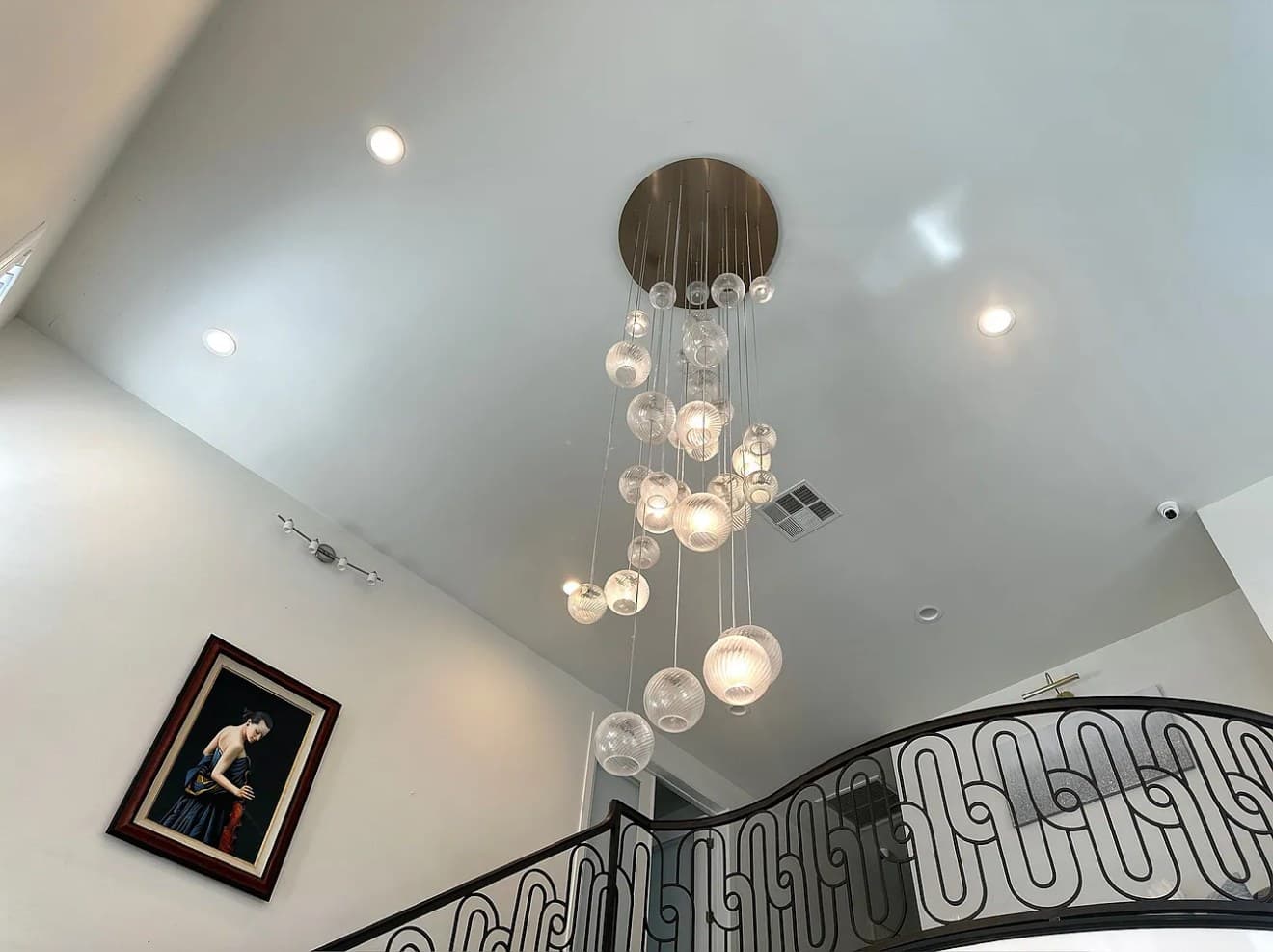 A glass-bubble chandelier hanging in a two-story entry, with a framed portrait on the wall and an iron stair rail below.