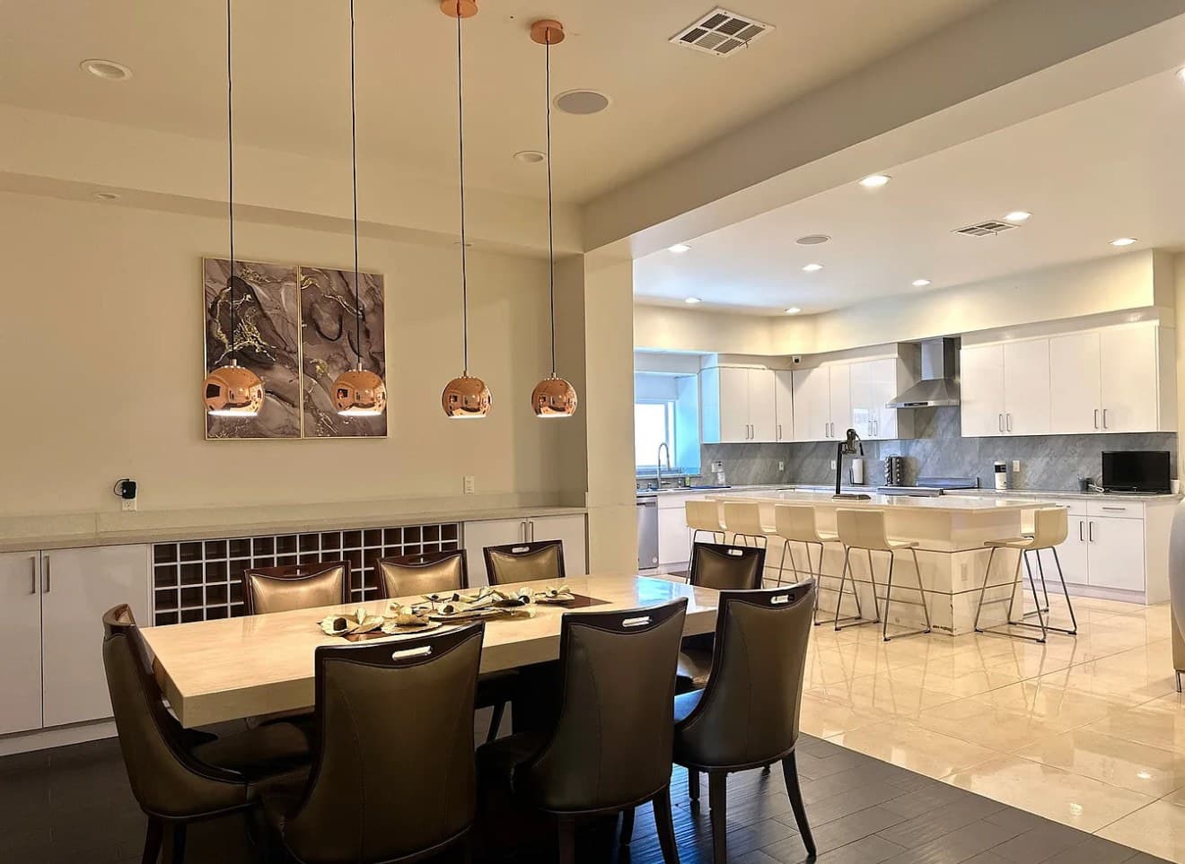 A dining table with leather chairs under copper pendant lights, with an open kitchen and bar stools behind it.